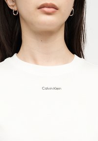 Νεαρό άτομο που φορά ασημένια κρίκους σκουλαρίκια και λευκό T-shirt Calvin Klein, με έμφαση στον λαιμό και το κάτω μέρος του προσώπου.