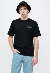 Μαύρο βαμβακερό t-shirt με κοντά μανίκια και στρογγυλή λαιμόκοψη, με μπλε λογότυπο «Dickies» στο αριστερό στήθος. Συνδυάζεται με ανοιχτόχρωμο τζιν και μαύρο καπέλο τύπου bucket.