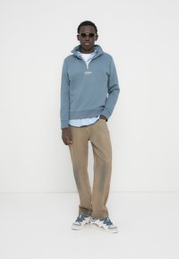 Φωτεινό γαλάζιο φούτερ με κουκούλα και μισό φερμουάρ, φορεμένο πάνω από ριγέ μπλε πουκάμισο, συνδυασμένο με ανοιχτό καφέ παντελόνι και αθλητικά παπούτσια.