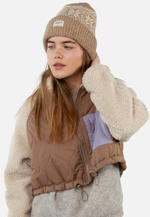 Brown quilted vest with a purple pocket and cream-colored shearling sleeves, worn over a gray sweater and paired with a patterned beige knit beanie.