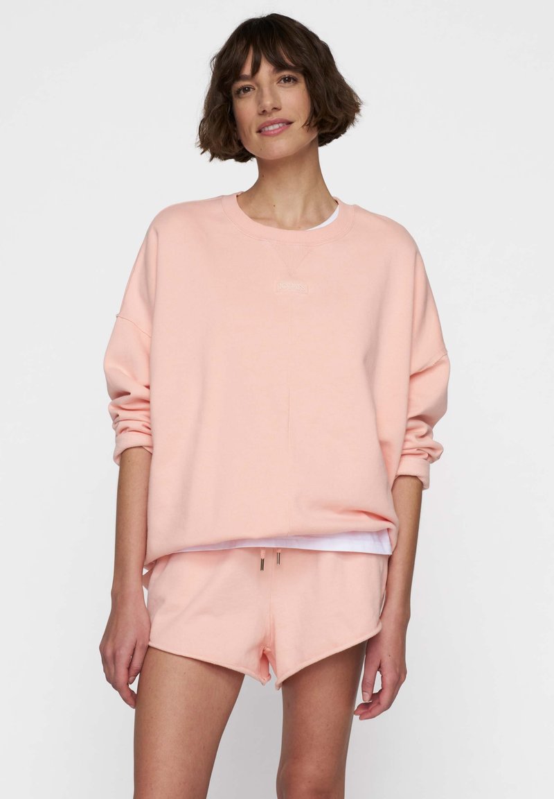Sudadera de crewneck rosa claro con hombros caídos, logo bordado y pantalones cortos a juego con cordón ajustable. Tela suave.