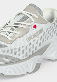 Άσπρο και γκρι sneaker Love Moschino με υφή πλεκτού, λευκά κορδόνια, χοντρή σόλα και μικρό κρεμαστό διακοσμητικό κορδονιού σε σχήμα καρδιάς.