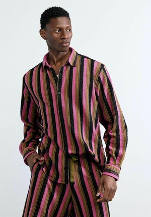 Man draagt een bijpassend gestreept shirt en broek in roze, zwart en bruin, staat met één hand in de zak en kijkt opzij.