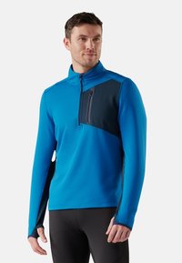 Suéter ligero de media cremallera en azul brillante, con un panel lateral azul oscuro y un bolsillo con cremallera en el pecho. Presenta agujeros para los pulgares en los puños.