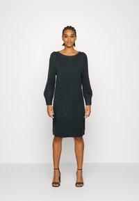 Abito lavorato a maglia verde scuro, con scollatura rotonda, maniche lunghe e silhouette dritta, abbinato a tacchi neri con laccetti.