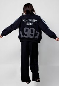 Must musternööriga jope "NORTHERN SOUL" ja "1987" tekstuurse tikandiga. Omab kontrastseid halli triipse varrukatel ja elastset vöökohta.