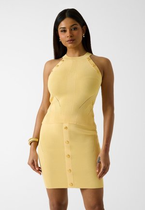 Femme portant une robe sans manches jaune pâle côtelée avec des détails de boutons sur les épaules et à l'avant, posant devant un fond uni.