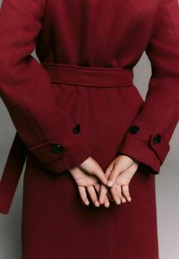 Maroon wool coat with a belted waist, large cuff buttons, and a textured finish, viewed from behind with hands clasped together.