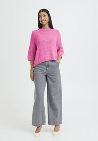 Femme debout portant un pull en peluche rose vif, un jean large gris et des chaussures blanches à bout pointu, souriante et regardant sur le côté.