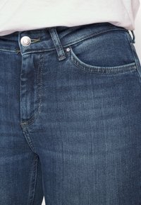 Jeans de denim en un lavado azul medio con un ligero desgastado. Presentan herrajes de botón plateado, un diseño tradicional de cinco bolsillos y detalles de costura.