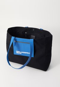 Svart canvas toteväska med blå handtag och en framficka med dragkedja. Har vit "KARL LAGERFELD JEANS" branding på fickan.