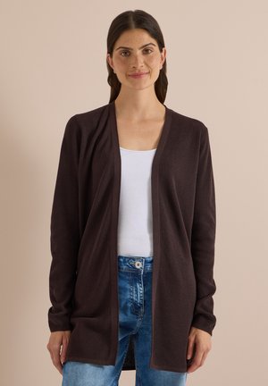 Femme aux cheveux foncés portant un cardigan marron foncé ouvert sur le devant, un haut blanc et un jean bleu, debout devant un fond beige uni.