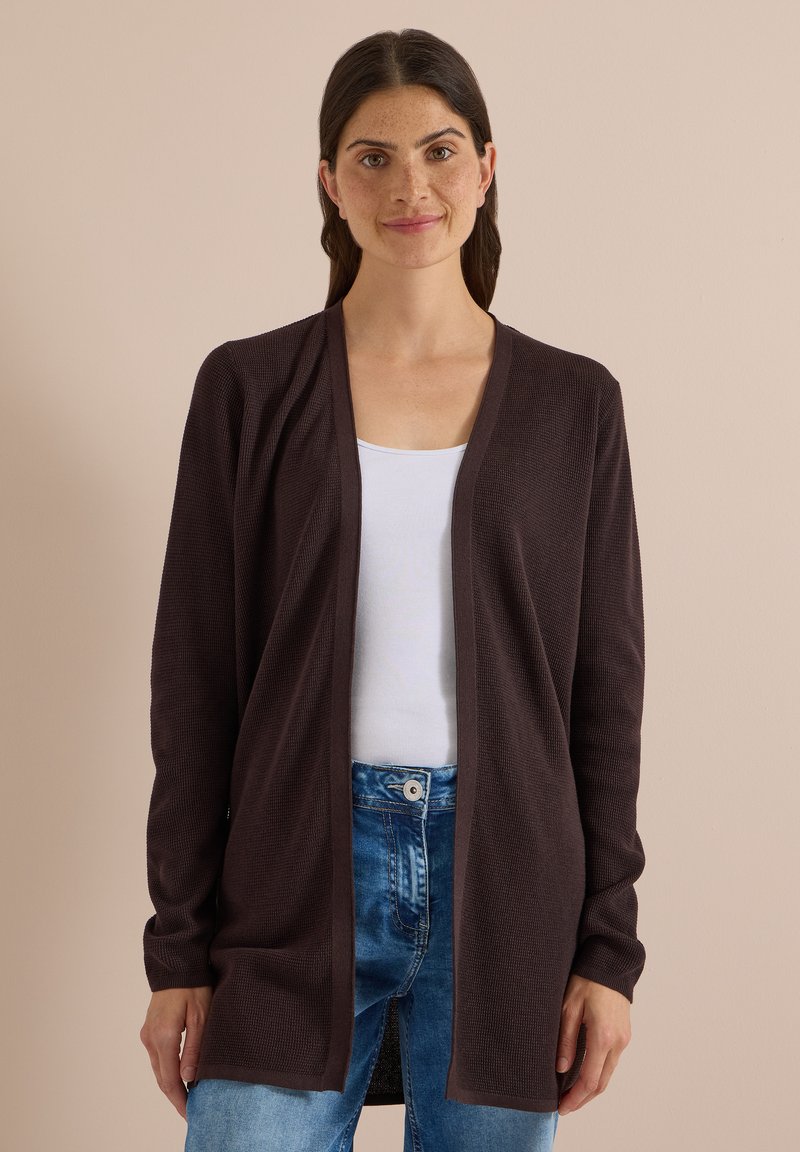 Femme aux cheveux foncés portant un cardigan marron foncé ouvert sur le devant, un haut blanc et un jean bleu, debout devant un fond beige uni.