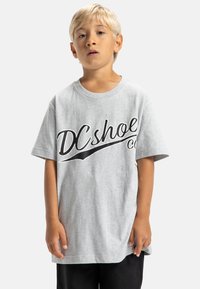 Šedé tričko s krátkým rukávem a černým logem "DCshoe". Bavlněný materiál, volný střih, kulatý výstřih. Model má na sobě černé kalhoty, ruce podél těla.