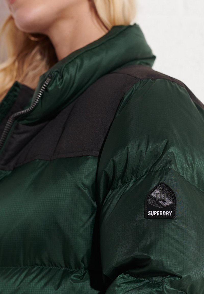 Kobieta w ciemnozielonej kurtce puchowej z czarnymi panelami na ramionach i naszywką z logo Superdry na górnej części rękawa w pobliżu zamka błyskawicznego.