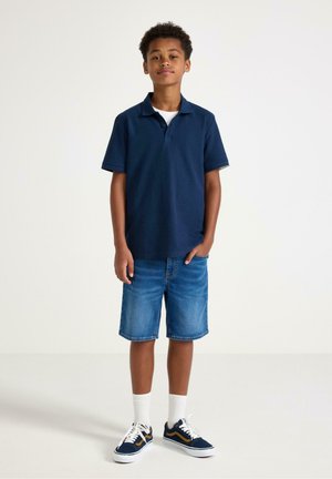 Dreng stående iført navyblå polo, denimshorts, hvide sokker og navy sneakers med orange striber på ensfarvet baggrund.