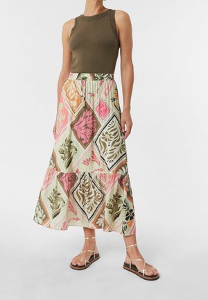 Woman wearing sleeveless olive top, long patterned skirt with floral diamond shapes, and strappy tan sandals against white background.