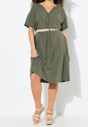 Woman wearing olive green belted V-neck dress with short sleeves and beige platform sandals, standing against white background.