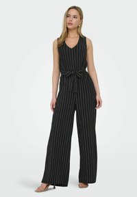 Mono negro sin mangas con rayas verticales blancas, escote en V, cinturón en la cintura y diseño de pierna ancha. Tejido suave y silueta minimalista.