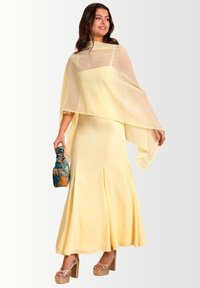 Geel satijnen jurk met een vloeiende transparante cape, uitlopende zoom en decoratieve stiksels. Gepaard met een gemusterde clutch en chunky hakken.