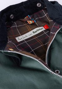 Πράσινη επένδυση με καρώ φόδρα σε καφέ, πορτοκαλί και λευκό. Διαθέτει ασημένιο φερμουάρ και ετικέτα που αναγράφει "Ο Paul Smith αγαπά τον Barbour."