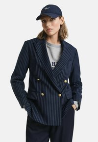 Blaser doppiopetto a righe navy con bottoni dorati, indossato sopra un maglione grigio, abbinato a un cappellino navy coordinato.