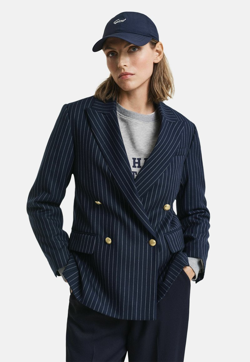 Blaser doppiopetto a righe navy con bottoni dorati, indossato sopra un maglione grigio, abbinato a un cappellino navy coordinato.