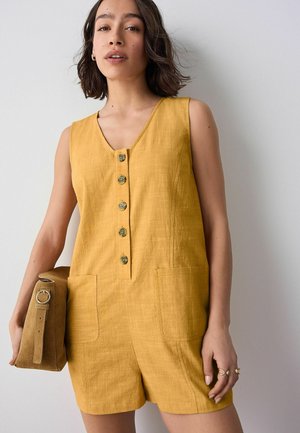 Yellow linen romper with a v-neckline, five button front, and two front pockets. Model holds a tan shoulder bag. Textured fabric.