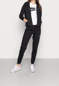 Felpa nera con zip e logo Nike bianco, abbinata a pantaloni jogger neri e sneakers Nike bianche/rosa. Tessuto liscio e vestibilità rilassata.
