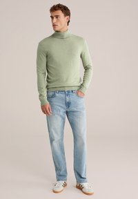 Hellgrüner Rollkragenpullover, schmal geschnitten, aus weichem Stoff. Kombiniert mit verwaschenen blauen Jeans und weißen Sneakers mit braunen Akzenten.