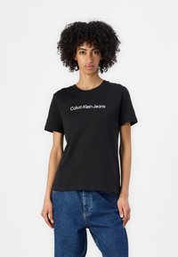 Melnā kokvilnas t-krekls ar īsām piedurknēm un apaļu kakla izgriezumu, ar baltu tekstu "Calvin Klein Jeans", kas drukāts pāri krūtīm.