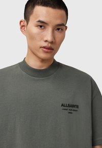 Camiseta de algodón verde oliva con cuello redondo y mangas cortas, que presenta un logo bordado de "ALLSAINTS" en el pecho. Textura suave.