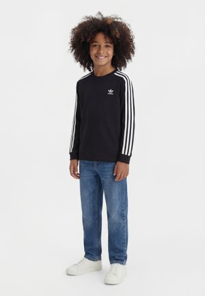 Enfant aux cheveux bouclés portant un t-shirt à manches longues Adidas noir, un jean bleu et des baskets blanches, debout et souriant sur fond blanc.