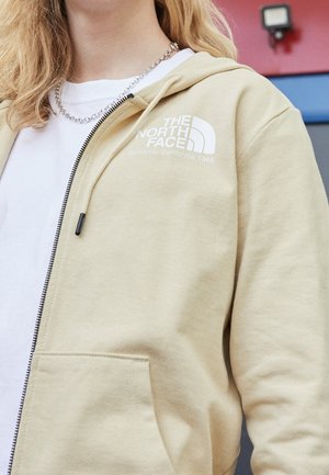 Person trägt eine beige Kapuzenjacke mit Reißverschluss und The North Face-Logo sowie ein weißes T-Shirt, silberne Ketten-Halskette, vor einem roten und blauen Hintergrund.
