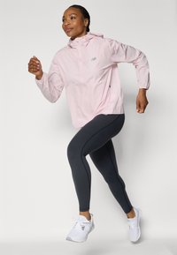 Light pink lightweight jacket with hood, features a zip front and reflective logo. Paired with dark gray leggings and white athletic shoes.