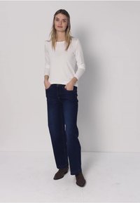 Frau steht mit Händen in den Taschen, trägt ein weißes Langarmshirt, dunkelblaue Jeans und braune Schuhe vor einem schlichten hellen Hintergrund.