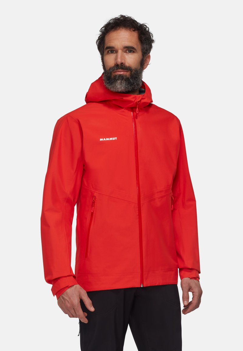 Giacca impermeabile rossa con cappuccio, dotata di tasche laterali con zip, polsini regolabili e un logo sul petto. Texture liscia.