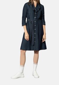 Denimmekko, jossa on röyhelökaulus, pitkät hihat ja vyötärölle sidottava nauha. Tumma sininen väri, jossa etupainikkeet ja liehuva hame. Pukeutunut valkoisiin tennareihin.