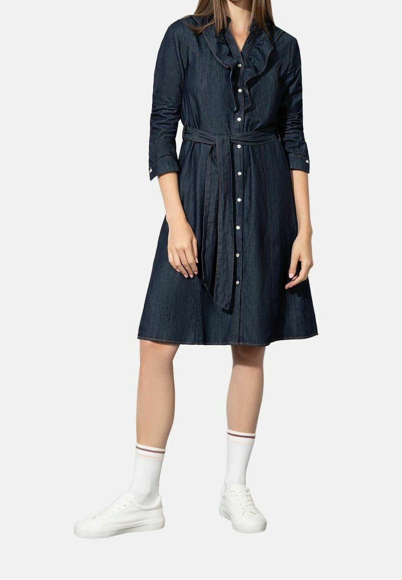 Denimmekko, jossa on röyhelökaulus, pitkät hihat ja vyötärölle sidottava nauha. Tumma sininen väri, jossa etupainikkeet ja liehuva hame. Pukeutunut valkoisiin tennareihin.