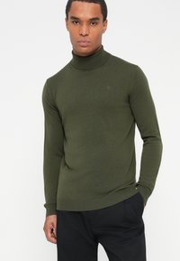Olivnozeleni pulover s turtleneck iz teksturirane pletenine, z dolgimi rokavi in subtilnim logotipom na prsih.