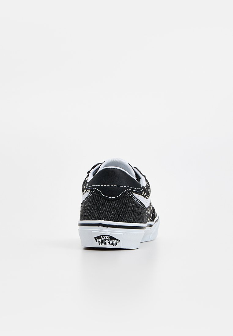 Zapatilla de lona negra con suela de goma blanca. Presenta costuras blancas y un logo destacado en el talón. Diseño clásico de corte bajo.