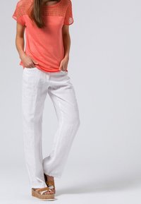 Korallfärgad topp med spetsliknande meshaxlar, kombinerad med vita linbyxor. Modellen bär ljusbruna plattformssandaler med remmar.