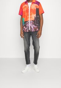 Chemise à manches courtes à boutons avec un imprimé vibrant de paysage urbain orange et violet, associée à un jean gris et des baskets blanches.