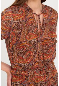 Ruskea kuviollinen mekko, jossa on paisley-kuvio punaisena, oranssina ja violetina. Mekossa on solmittava kaulus ja tupsuakselit, sekä laskostetut hihat.