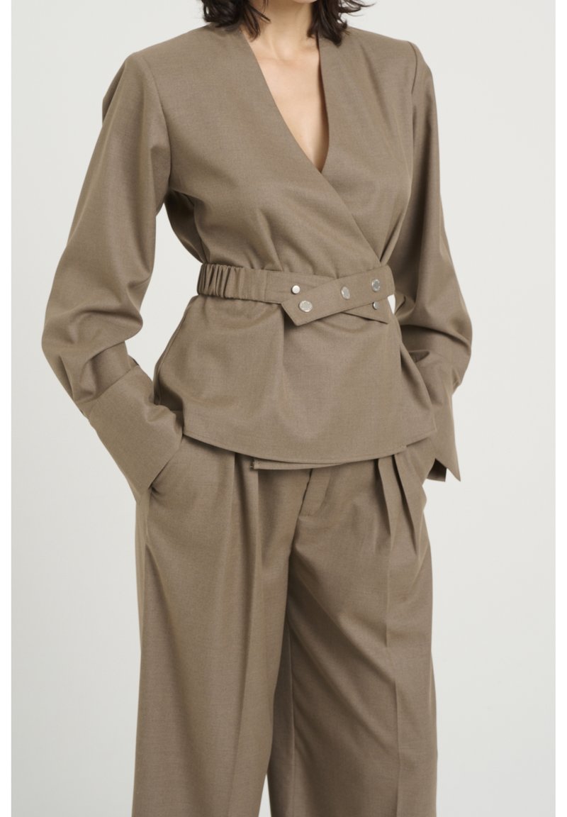 Haut marron de style portefeuille avec manches longues, doté d'une taille ceinturée et de fermetures à pression argentées. Tissu texturé avec une coupe ajustée.