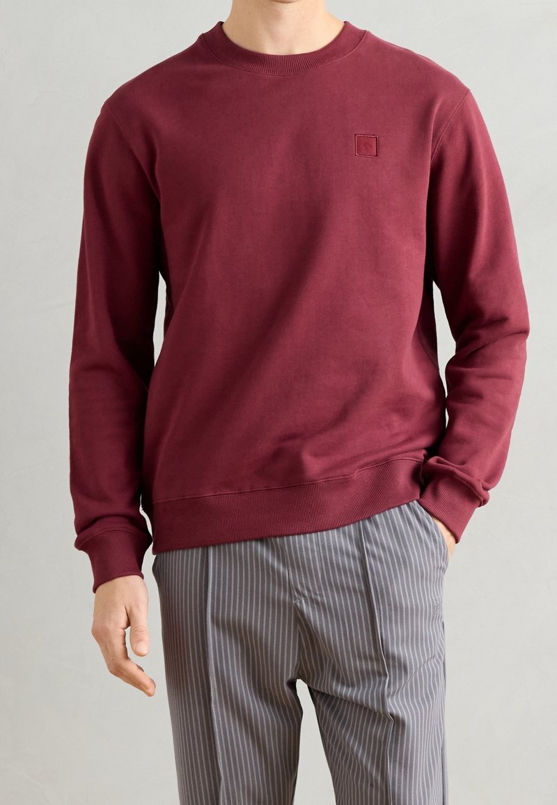 Homme portant un sweat-shirt bordeaux uni et un pantalon gris à fines rayures, debout avec une main dans la poche devant un fond neutre.