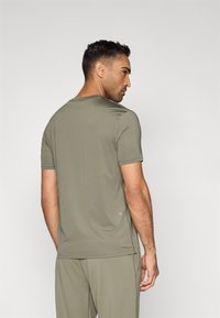 T-shirt de manga curta, gola redonda, em verde-oliva, feita de tecido suave. Apresenta um detalhe discreto com o logo perto da bainha e um corte ajustado.