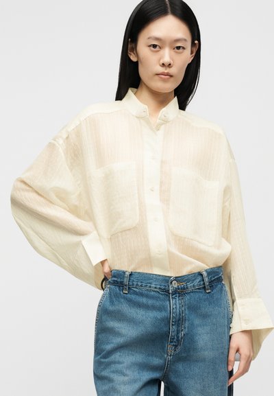 Femme aux longs cheveux noirs portant une chemise crème transparente surdimensionnée boutonnée et un jean bleu taille haute, debout devant un fond neutre.