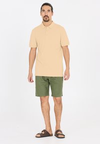 Vaaleanbeige-poolopaita klassisella kauluksella, yhdistettynä vihreisiin shortseihin. Ruskeissa sandaaleissa on solkiyksityiskohta ja kontrastinen pohja.