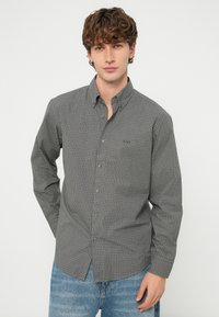 Fiatal férfi szürke kockás gombos ingben és kék farmerral, egy kézzel a háta mögött, egyszerű háttér előtt áll.